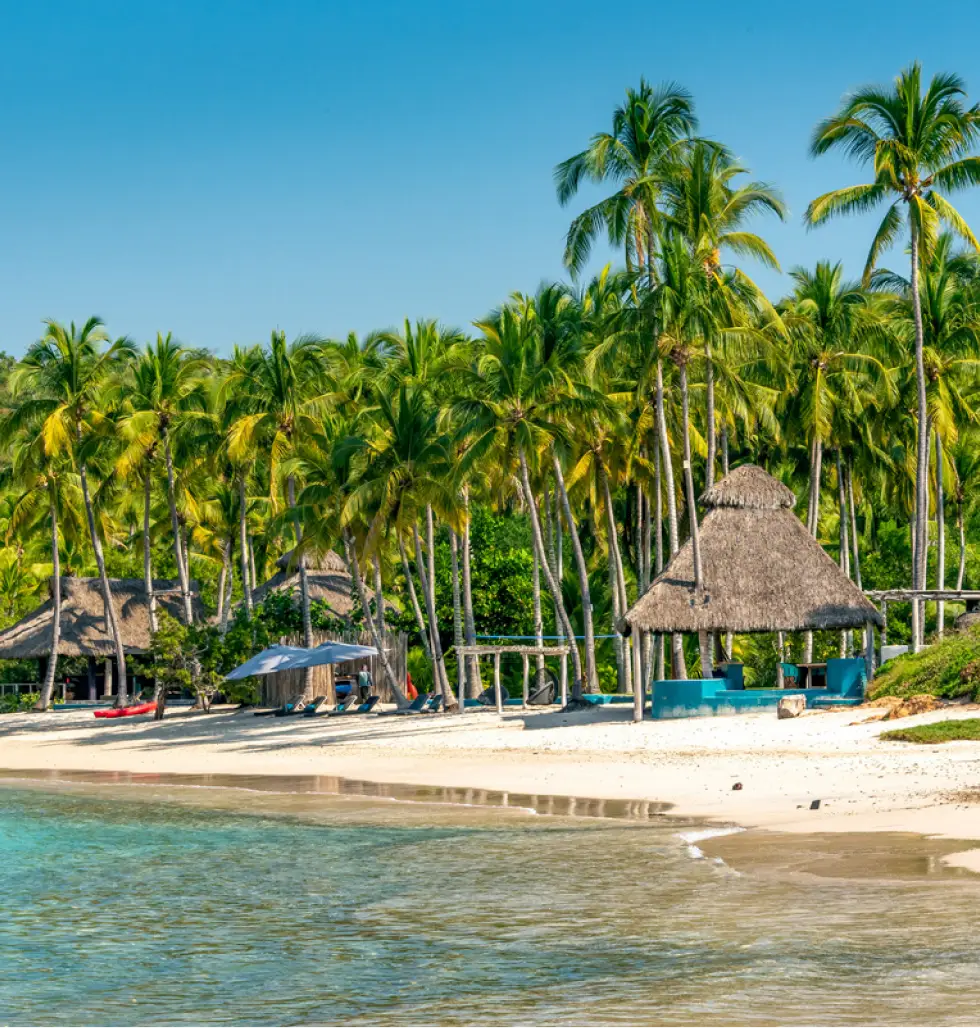 Playa tropical con palmeras, cabañas de paja y sillas de playa junto al agua cristalina